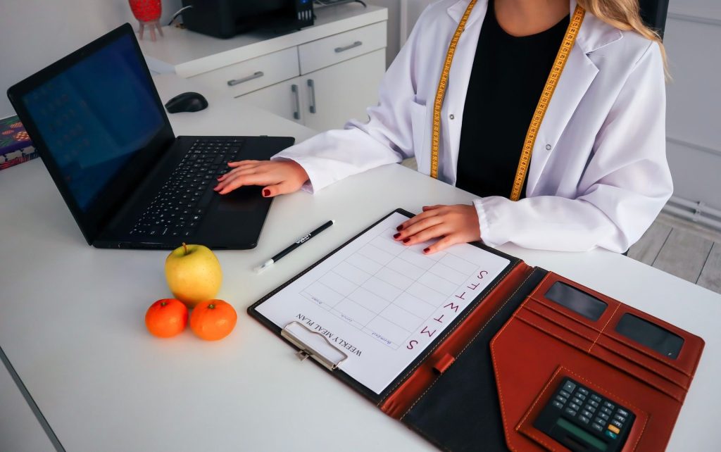 Ernährungsberaterin plant am Schreibtisch mit Laptop und Wochenplan die Ernährung eines Klienten